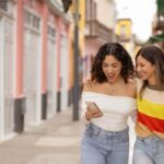 アメリカのカラフルな街並みを背景に、スマホを見ながら笑顔で歩く2人の若い女性。肩を寄せ合いながら仲良くショッピングストリートを散策している様子が写っている。