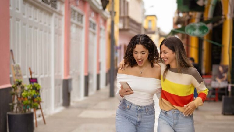 アメリカのカラフルな街並みを背景に、スマホを見ながら笑顔で歩く2人の若い女性。肩を寄せ合いながら仲良くショッピングストリートを散策している様子が写っている。