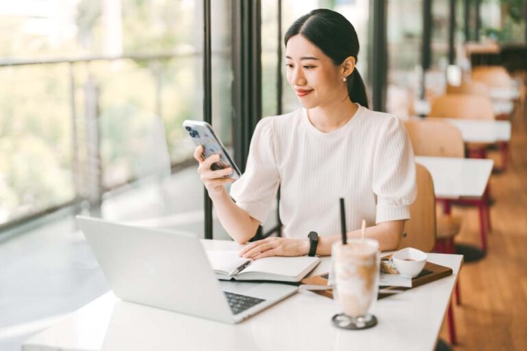 カフェの窓際でスマートフォンとノートパソコンを使いながら作業するアジア系女性。ドリンクを片手に、仕事や勉強に集中している穏やかな時間。