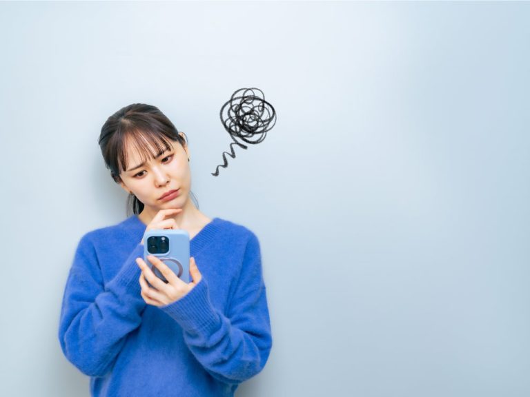 青いセーターを着た女性がスマートフォンを見て困惑した表情をしている様子。頭の上に混乱を表す黒い渦巻きが描かれている。