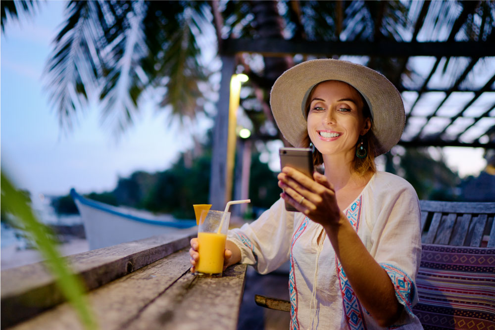 屋外のリゾートで帽子をかぶり、スマートフォンを操作しながら飲み物を楽しんでいる女性