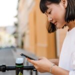 スマートフォンでアプリを操作する若い女性。電動キックボードの前で立ち止まり、街中で移動手段を確認している様子。