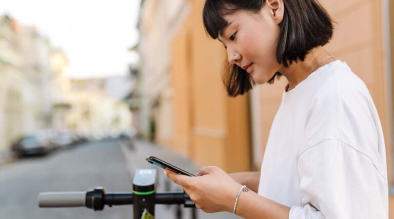 スマートフォンでアプリを操作する若い女性。電動キックボードの前で立ち止まり、街中で移動手段を確認している様子。