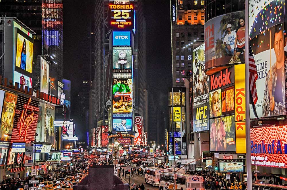 タイムズスクエアの夜景。きらびやかなネオンサインや広告が街を明るく照らし、賑やかな通りに多くの車や観光客が集まっている。