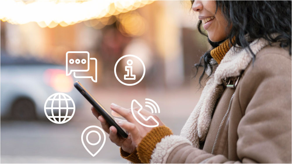  街中でスマートフォンを操作する女性の横顔。背景はぼかされ、温かみのあるライトが灯る。画面上に「メッセージ」、「情報」、「地球儀」、「位置情報」、「電話」のアイコンがオーバーレイされ、コミュニケーションや情報の連携をイメージしている。