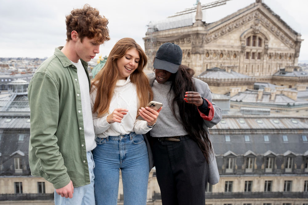 屋外の屋上でスマートフォンを一緒に見ている3人の友人たち。背景には歴史的な建物が広がり、楽しく会話しながら笑顔を浮かべている様子が写されている。