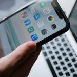 手に持たれたスマートフォンの画面。InstagramやFacebookなどのアプリが表示され、背景にはノートパソコンのキーボードが写っている。