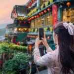 台湾・九份の美しい街並みを背景に、スマートフォンで写真を撮る長い髪の女性。夕暮れの空と赤い提灯が彩りを添えている。