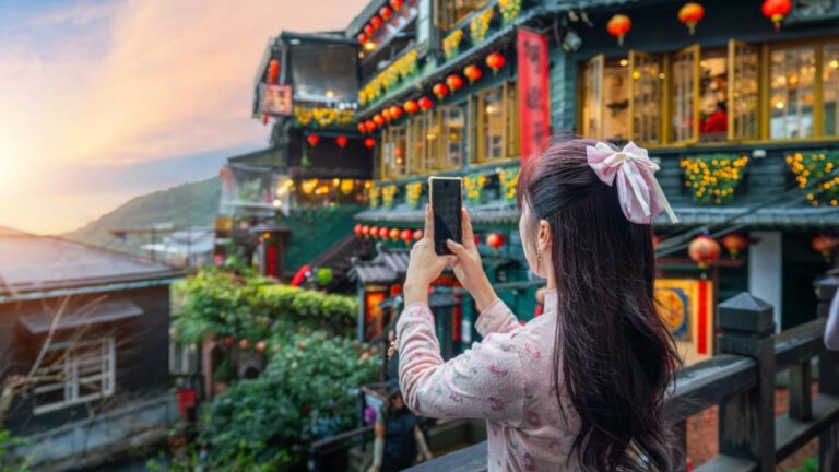 台湾・九份の美しい街並みを背景に、スマートフォンで写真を撮る長い髪の女性。夕暮れの空と赤い提灯が彩りを添えている。