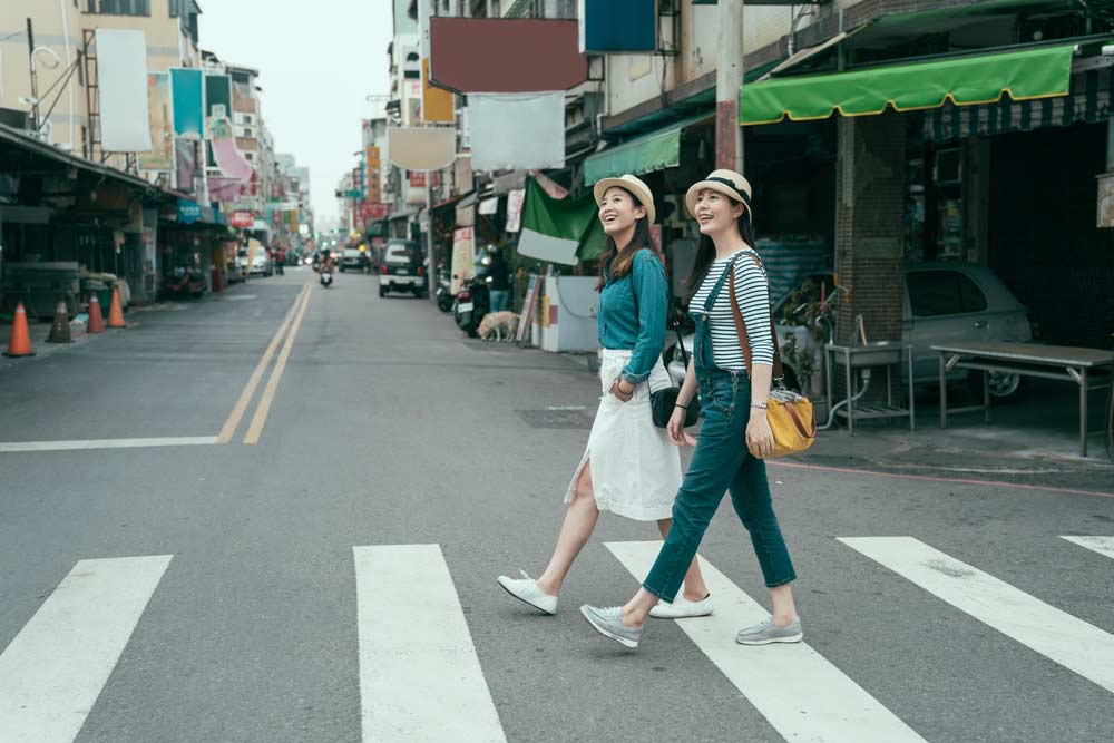 台湾のローカルな街並みを背景に、横断歩道を楽しそうに歩く2人の若い女性旅行者。観光や街歩きの楽しさを伝えるシーンに最適。