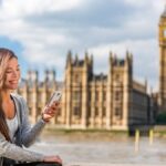 ロンドンのビッグベンと議会議事堂を背景に、スマートフォンを見ながら微笑む女性。