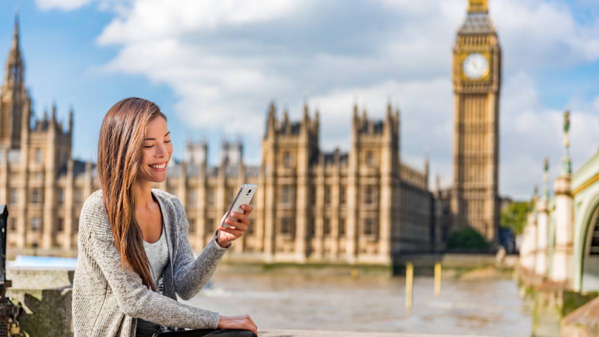 ロンドンのビッグベンと議会議事堂を背景に、スマートフォンを見ながら微笑む女性。