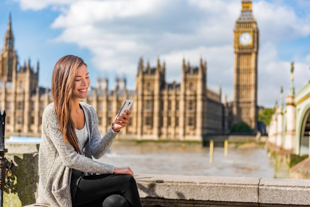 ロンドンのビッグベンと議会議事堂を背景に、スマートフォンを見ながら微笑む女性。