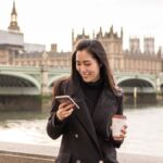 イギリス・ロンドンのビッグベンを背景に、スマートフォンを見ながらコーヒーを持つ女性