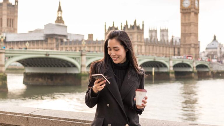 イギリス・ロンドンのビッグベンを背景に、スマートフォンを見ながらコーヒーを持つ女性