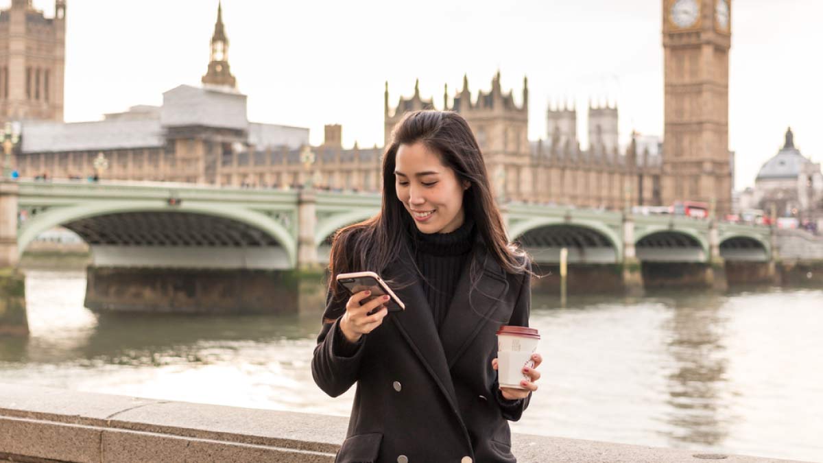 イギリス・ロンドンのビッグベンを背景に、スマートフォンを見ながらコーヒーを持つ女性