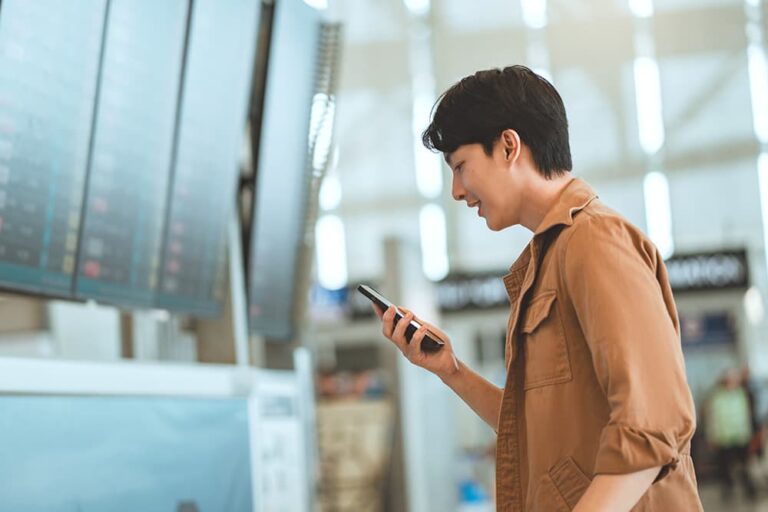 空港の案内板の前でスマホを操作し、海外旅行用eSIMに切り替える男性