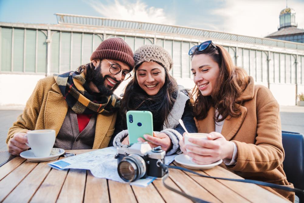 屋外のカフェでテーブルを囲む3人の友人がスマートフォンを見て笑顔で楽しんでいる様子。テーブルにはカメラと地図が置かれ、旅行中のワンシーンを表現している。