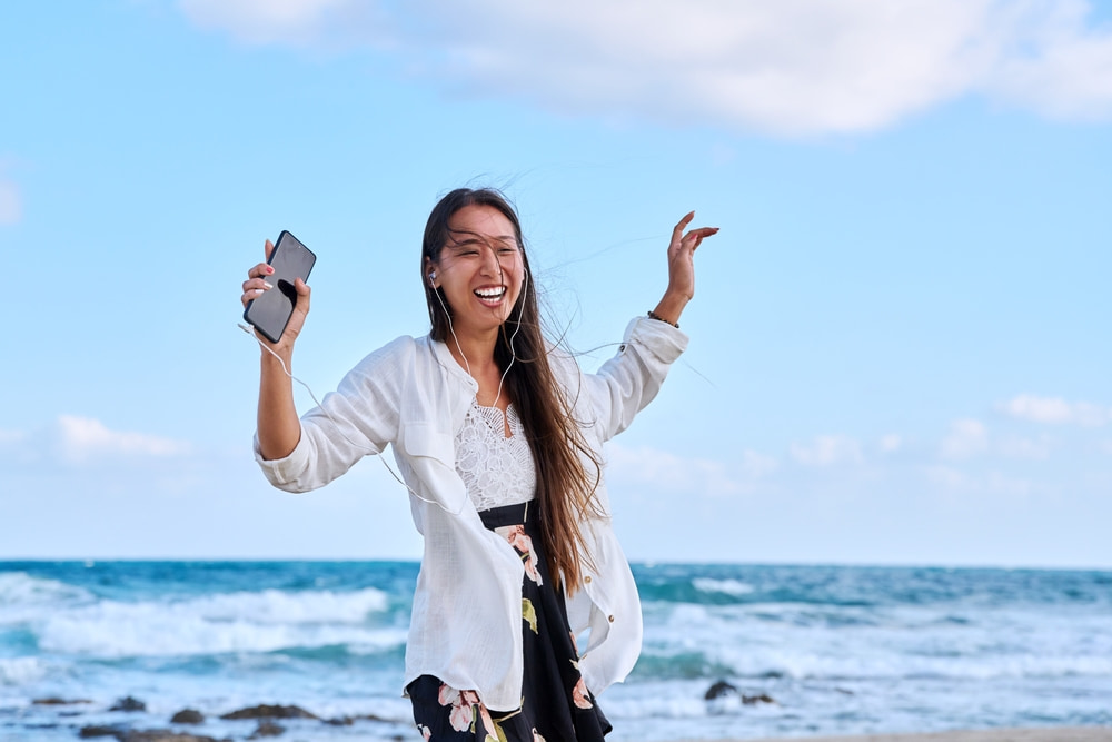 海辺でイヤホンをつけて音楽を聴きながらスマートフォンを手に持ち、楽しそうに踊る女性。