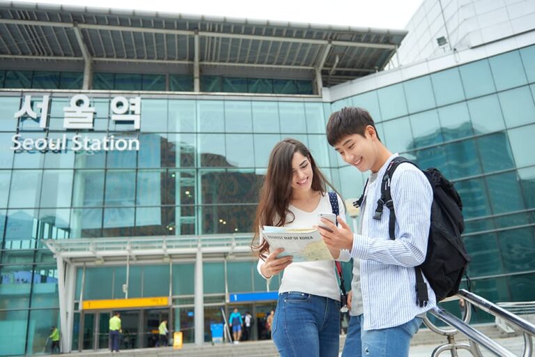 韓国旅行での通信費(Wi-FiやSIM)の利用シーンをイメージした、ソウル駅前でスマートフォンを使って観光情報を調べる男女の旅行者
