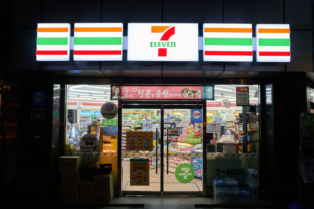 韓国・ソウルにある7-Eleven（セブンイレブン）の店舗外観。夜に明るく照らされた店内には、スナックや飲料、日用品が並ぶ。店頭にはATMやプロモーション広告が掲示されている。