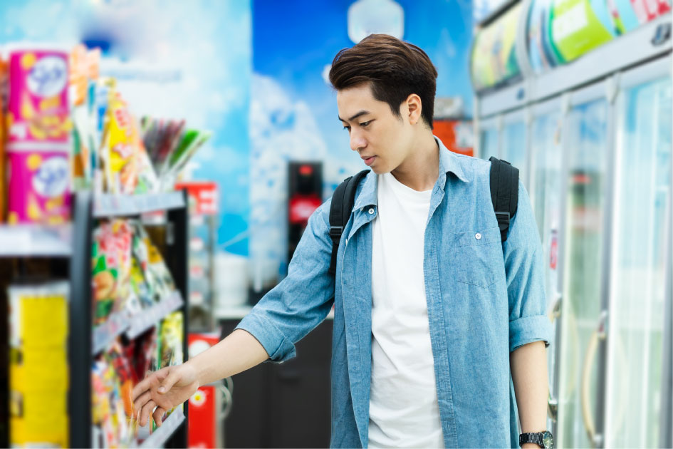 韓国のコンビニで商品を選ぶ若い男性。デニムシャツを着てバックパックを背負い、棚に並ぶスナックを手に取ろうとしている。