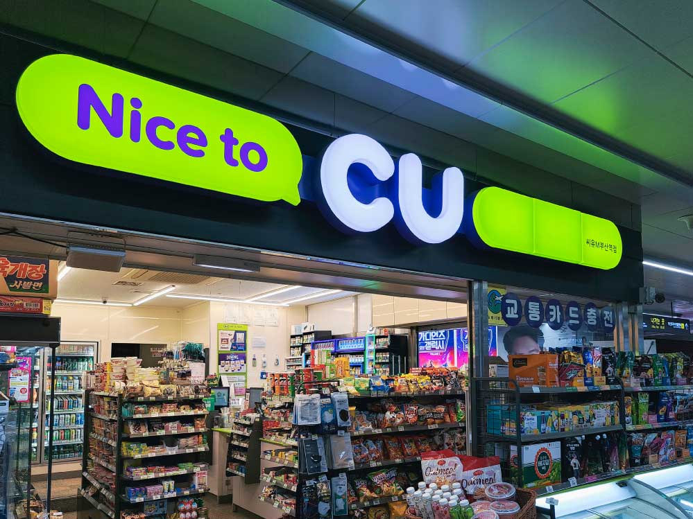 韓国・釜山にあるCU（シーユー）の店舗外観。緑と紫の看板が特徴的で、「Nice to CU」のスローガンが掲げられている。店内にはスナック、飲料、日用品などが陳列され、交通カードのチャージサービスも提供されている。