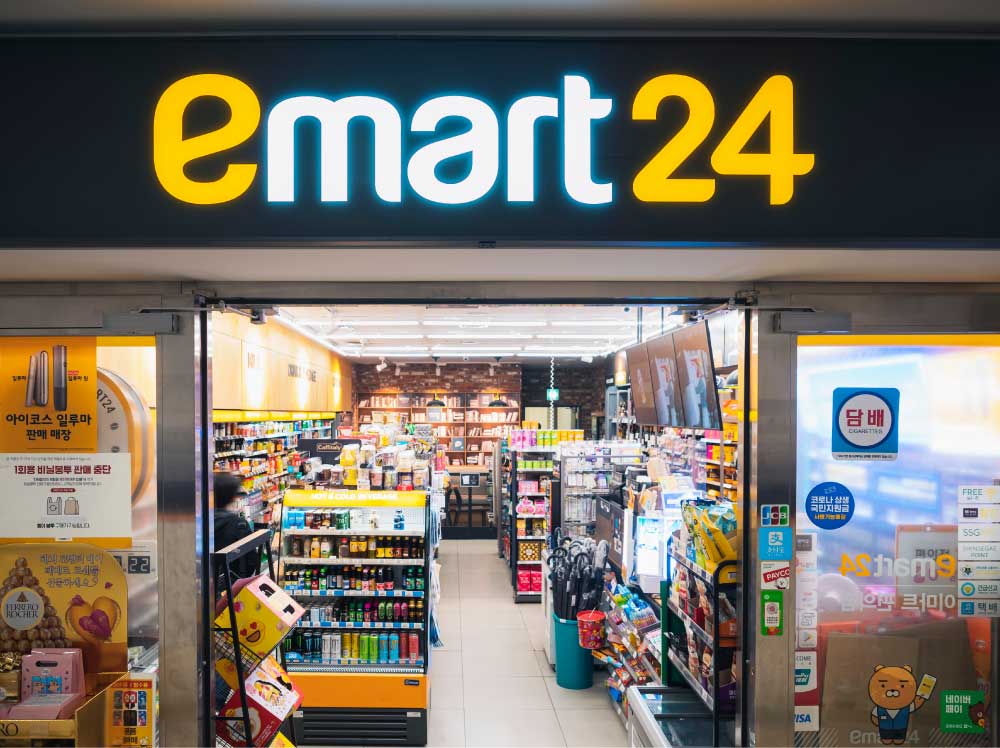 韓国・釜山にあるemart24（イーマート24）の店舗外観。黒と黄色の看板が特徴的で、店内には飲料、スナック、お菓子、生活用品が揃っている。入口にはタバコ販売の案内やキャラクターグッズの広告が貼られている。