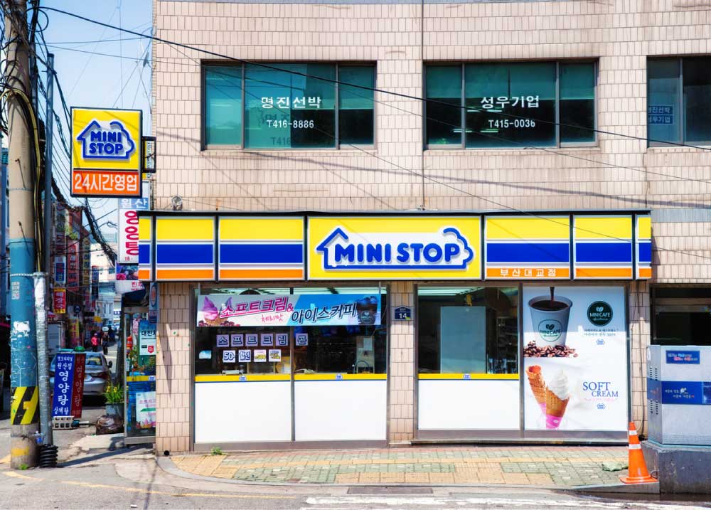 韓国・釜山にあるMINI STOP（ミニストップ）の店舗外観。黄色と青の看板が特徴的で、「24時間営業」の表示がある。店の窓にはソフトクリームやアイスコーヒーの広告が貼られている。
