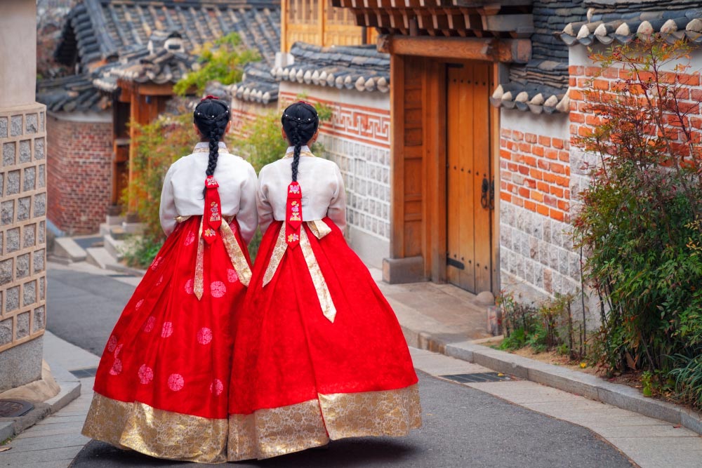 韓国の伝統的な韓屋の街並みを歩く、赤と金色の韓服（ハンボク）を着た2人の女性の後ろ姿。歴史的な雰囲気が漂う静かな路地。