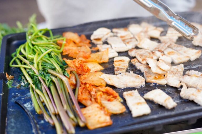 韓国の人気焼肉料理「ティッコギ」（薄切り豚肉の焼肉）。鉄板の上で香ばしく焼き上げられた豚肉に、キムチやニンニク、水パセリを添えて楽しむ。