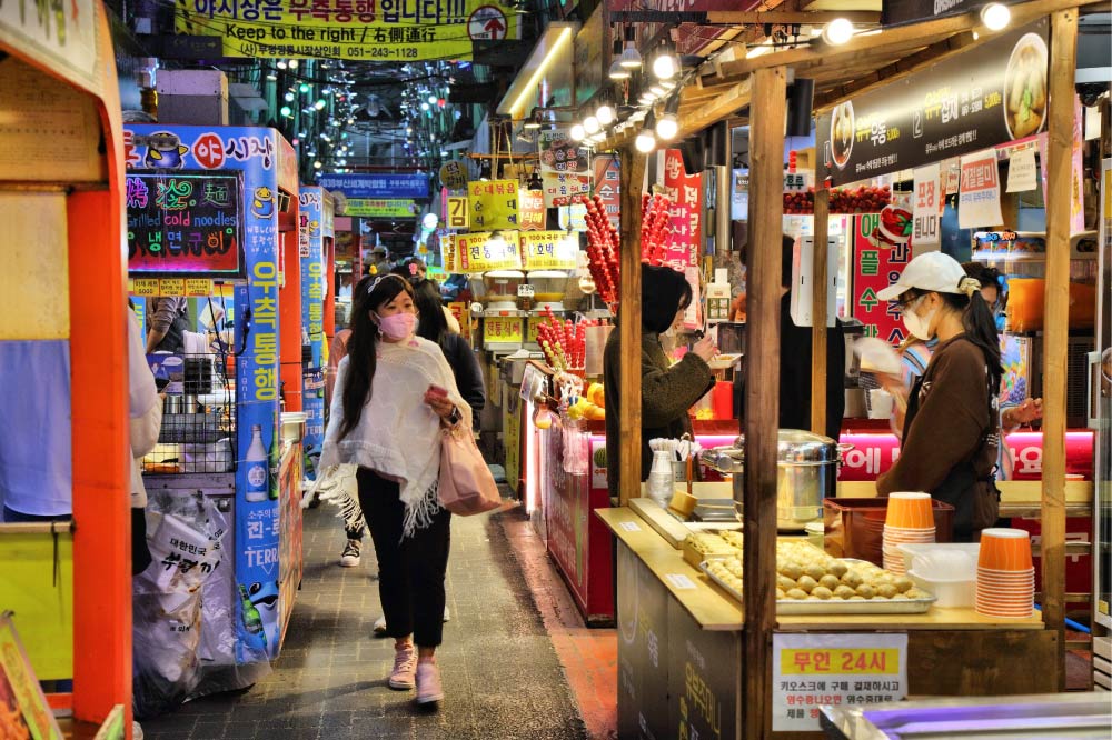 韓国・釜山の富平カントン市場の夜市の様子。色鮮やかな看板や提灯が並び、活気あふれる雰囲気の中で、観光客や地元の人々が屋台グルメを楽しんでいる。