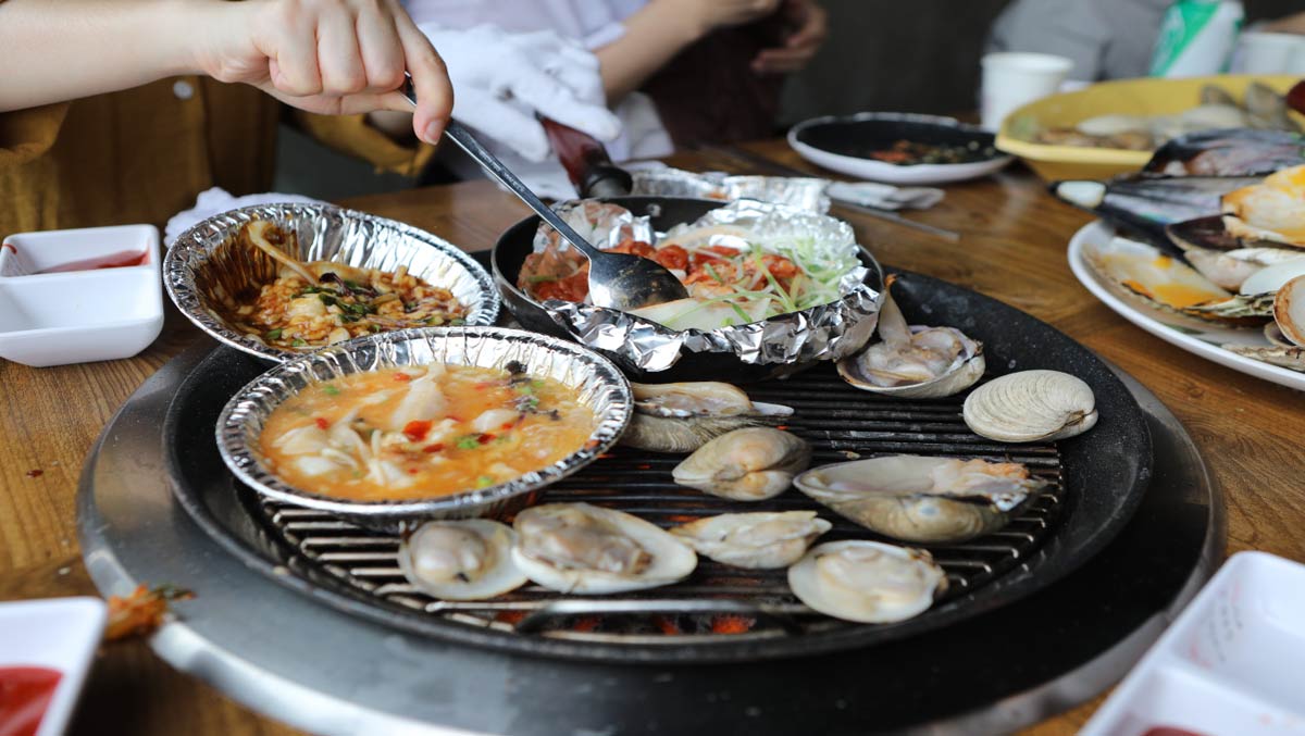 韓国の海鮮バーベキューの様子。網の上で新鮮な貝類やホイル焼きの海鮮料理が焼かれている。