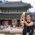 ソウルの景福宮（キョンボックン）前で自撮りをする女性観光客。伝統的な建築と背景に広がる現代的な高層ビルが対照的。