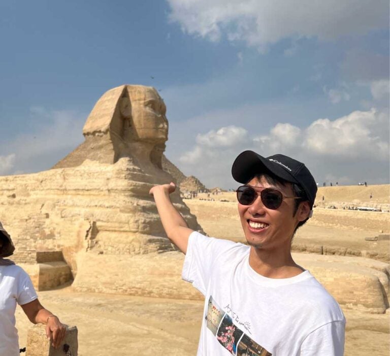 エジプト・ギザのスフィンクス前で写真を撮る旅行者。晴れ渡る青空の下、スフィンクスに手を添えるポーズをとり、楽しそうな笑顔を浮かべている。