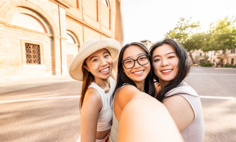 ヨーロッパの街角でセルフィーを撮る3人の若い女性旅行者。明るい日差しのもと、笑顔で楽しそうな表情を見せており、旅行中の友情や楽しい思い出を感じさせる1枚。