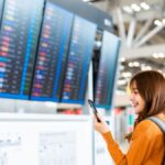空港の出発案内板の前でスマートフォンを見ながら微笑むバックパック姿の女性旅行者。出発前のワクワク感と旅への期待が感じられるシーン。