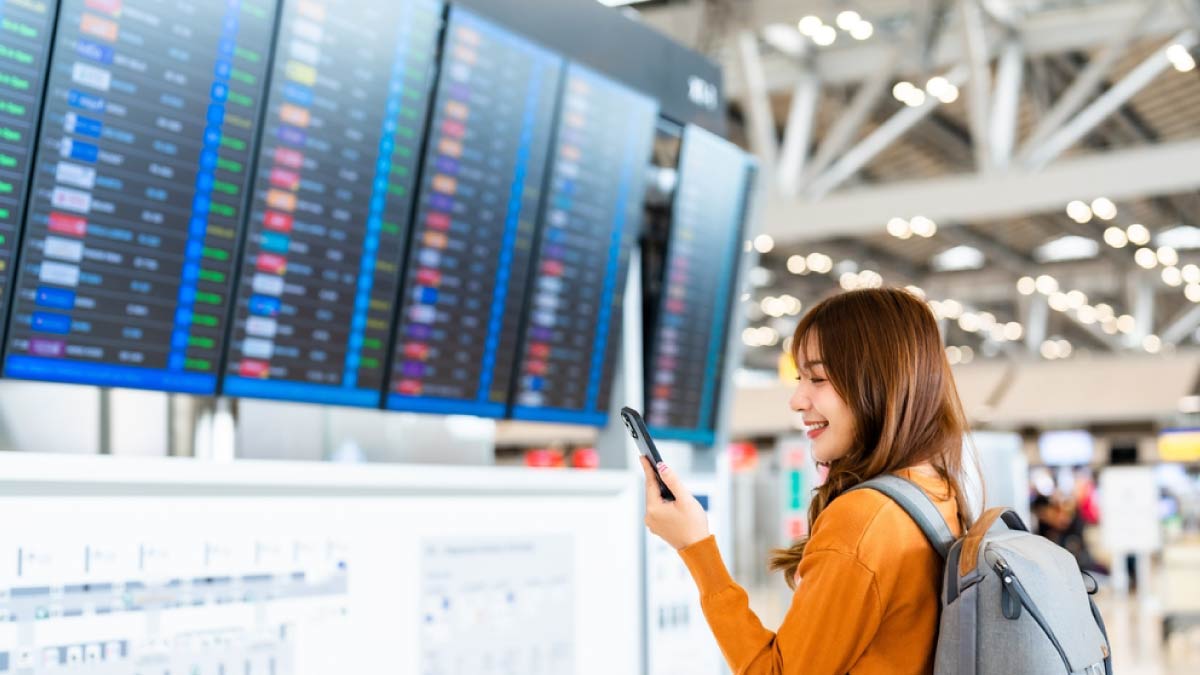 空港の出発案内板の前でスマートフォンを見ながら微笑むバックパック姿の女性旅行者。出発前のワクワク感と旅への期待が感じられるシーン。