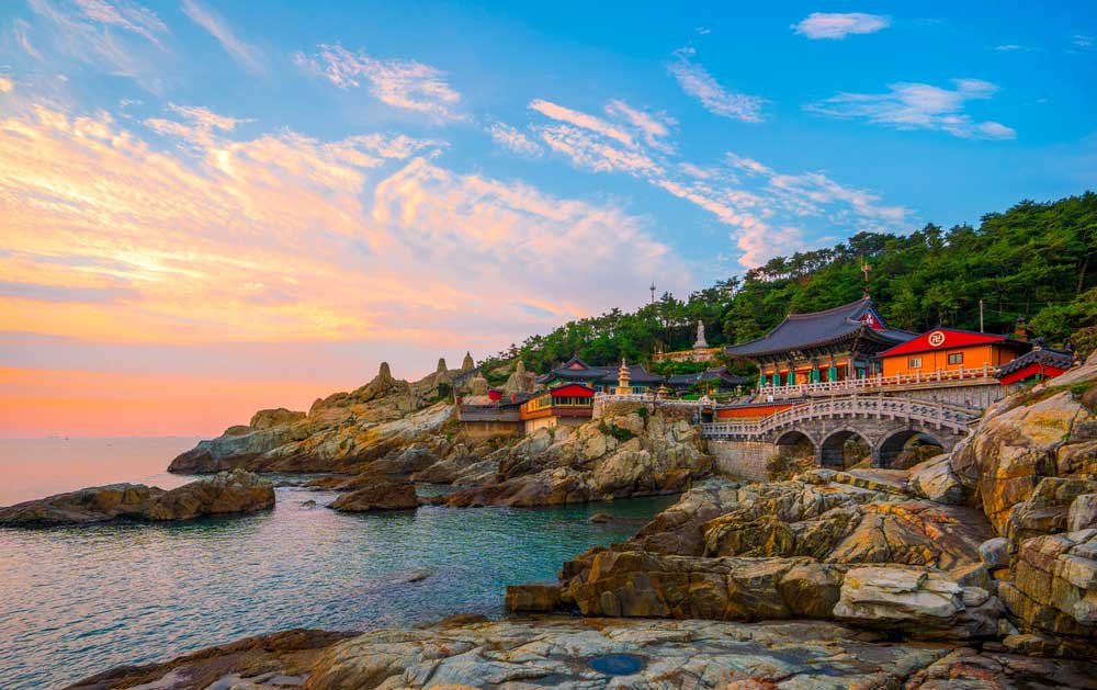 韓国・釜山にある海沿いの寺院、海東龍宮寺(ヘドンヨングンサ)。夕焼けの空と海が広がる中、岩場に佇む美しい伝統建築が印象的で、観光スポットとしても人気の絶景スポット。