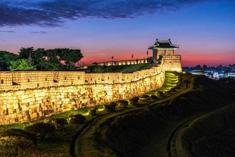 韓国・水原にある華城（ファソン）の夜景。ライトアップされた城壁と、夕焼けに染まる空が幻想的な雰囲気を演出している歴史的な風景。