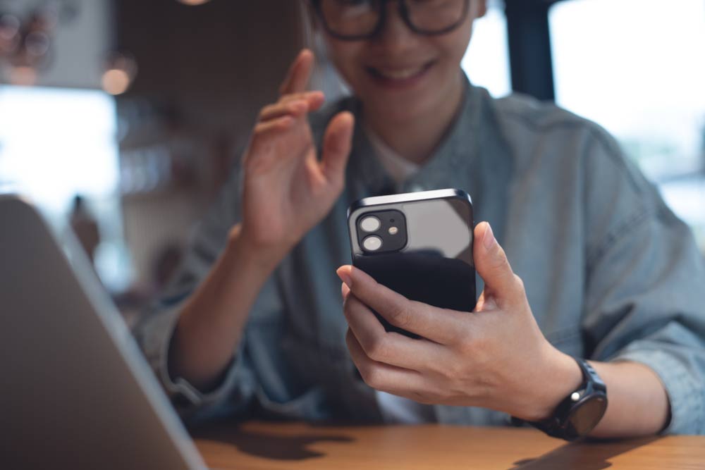カフェでスマートフォンを使ってビデオ通話を楽しむアジア系の女性。笑顔で手を振りながら会話するリラックスした雰囲気のワンシーン。