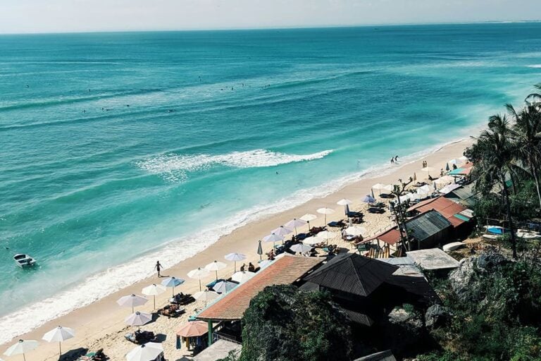 8月のバリ島・デンパサールで、白砂のビーチ沿いに並ぶパラソルと海を楽しむ観光客の様子を紹介する写真