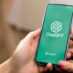 女性が手に持つスマートフォンの画面に表示されたChatGPTのアプリ起動画面。緑色の背景にChatGPTとOpenAIのロゴが見える。