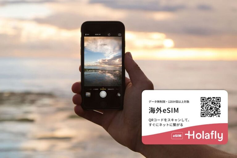 海外の海辺でスマートフォンを使って風景を撮影する様子と、HolaflyのeSIM広告を組み合わせた写真。旅先でもネットが使える便利さを伝えるビジュアル