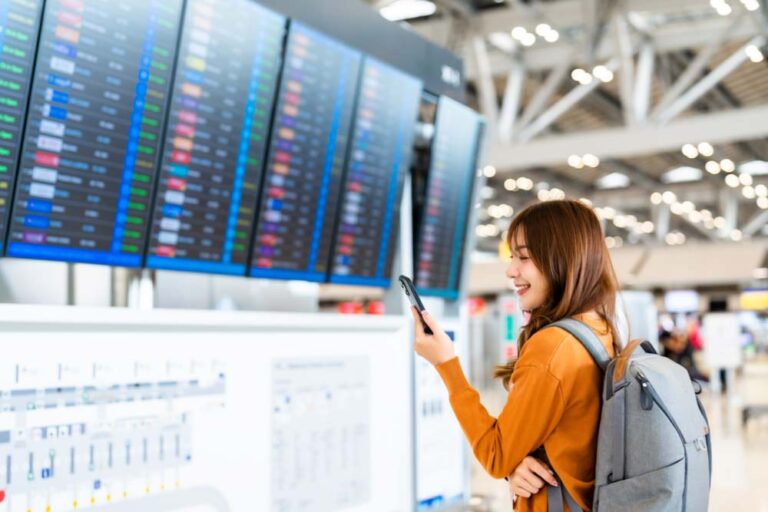 空港の出発案内板の前でスマートフォンを見ながら微笑むバックパック姿の女性旅行者。出発前のワクワク感と旅への期待が感じられるシーン。