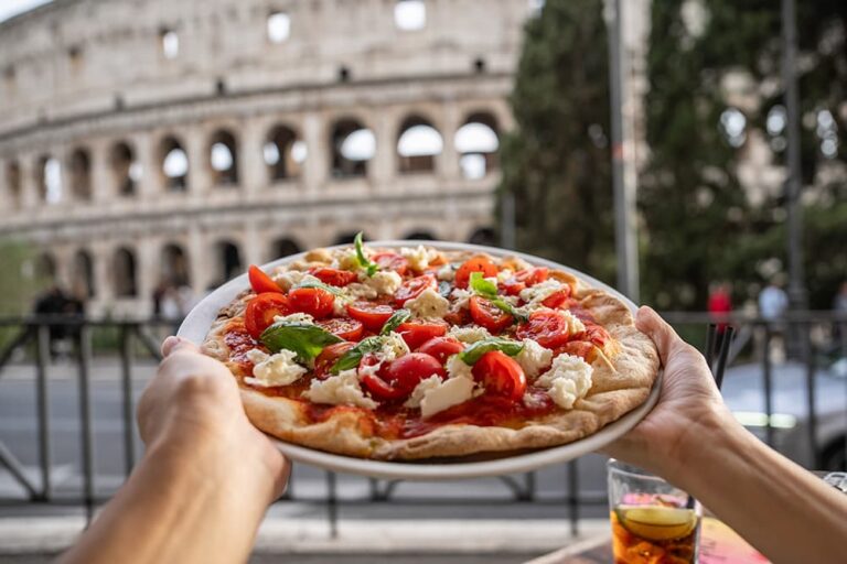ローマのコロッセオを背景にトマトとモッツァレラのピザを掲げるイタリア美食の一枚