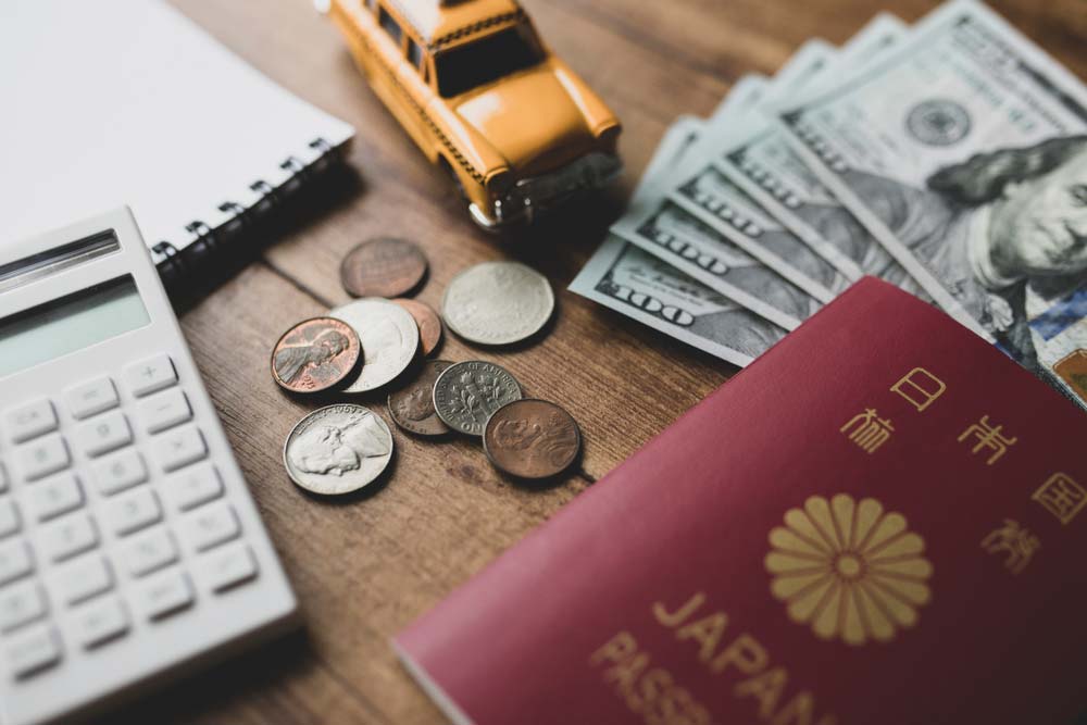 日本のパスポート、アメリカドル紙幣と硬貨、電卓、ノート、ミニチュアのタクシーが並ぶ木製テーブル。海外旅行前の予算計画や資金準備をイメージした構成。