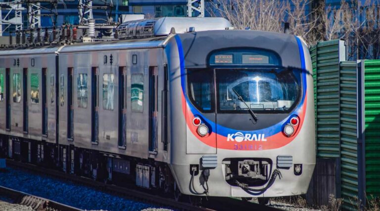 韓国・KORAIL(コレール)の通勤電車が線路を走行している様子。青と赤のラインが特徴的な車両で、都市部の防音壁に囲まれた区間を進む姿が写されている。