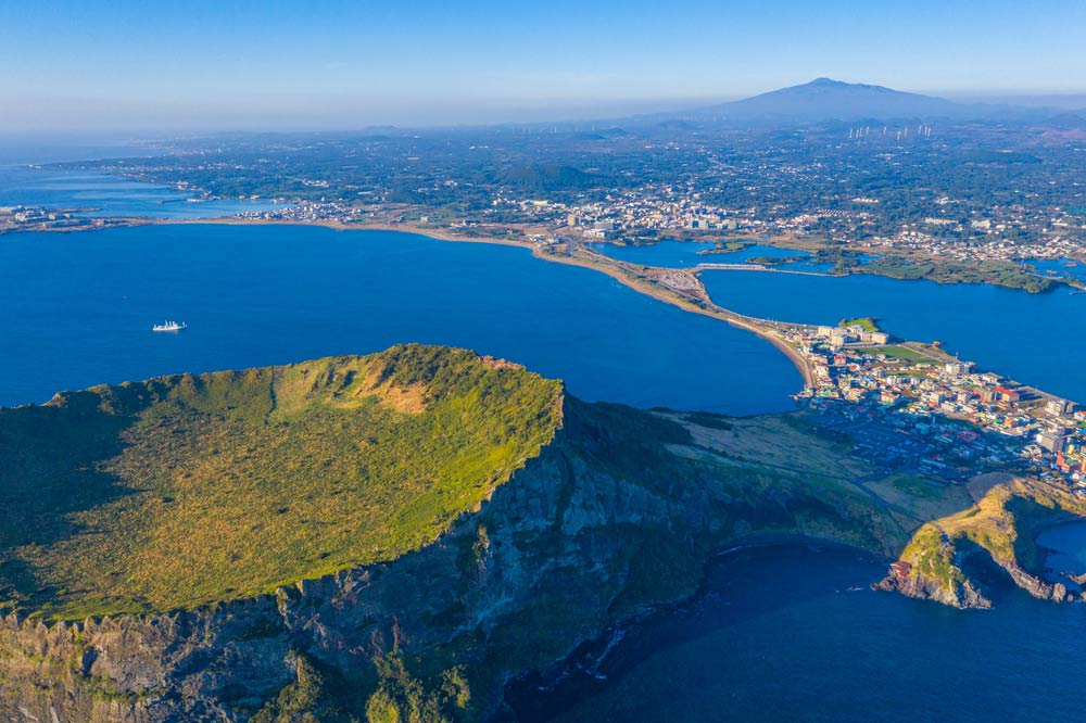 韓国・済州島の城山日出峰を上空から望む風景。噴火口の縁に沿って広がる緑と青い海、遠くにそびえる漢拏山が島の自然の雄大さを物語る。