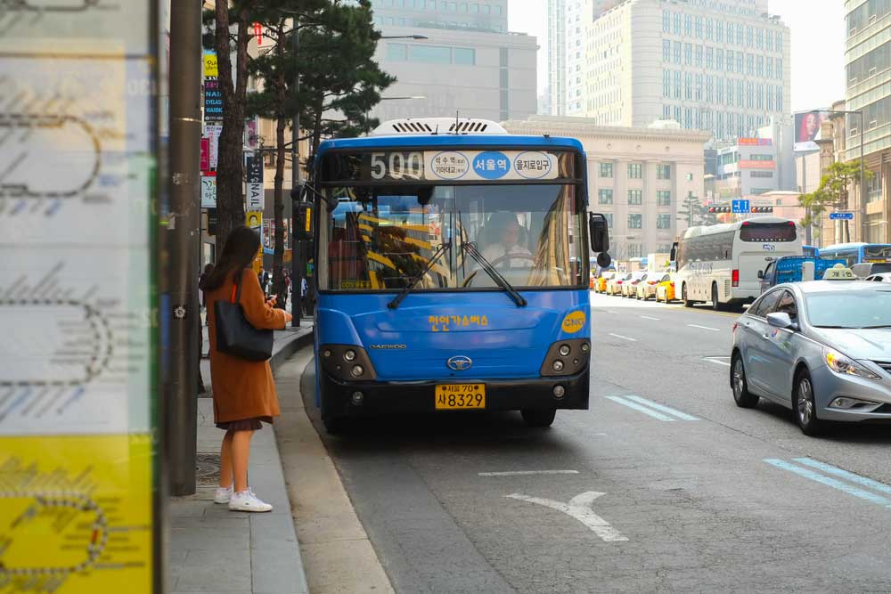 韓国・ソウルの市内を走る青い500番バス。乗車を待つ女性と、背景には高層ビルが立ち並ぶ都心の様子が映されている。